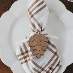 May include: A white plate with a scalloped edge, a brown and white checkered napkin, and a wooden pine cone napkin ring. The napkin ring is tied with a white ribbon. A gold fork and spoon are on the right side of the plate.
