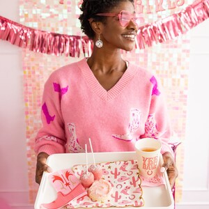 K&ouml;nnte beinhalten: Ein rosa Pullover mit Cowboy-Stiefel-Muster, ein wei&szlig;es Tablett mit rosa und goldenen Servietten, zwei rosa Kuchen am Stiel, ein rosa glasierter Keks und eine rote Tasse mit dem Text "Yee Haw" darauf.
