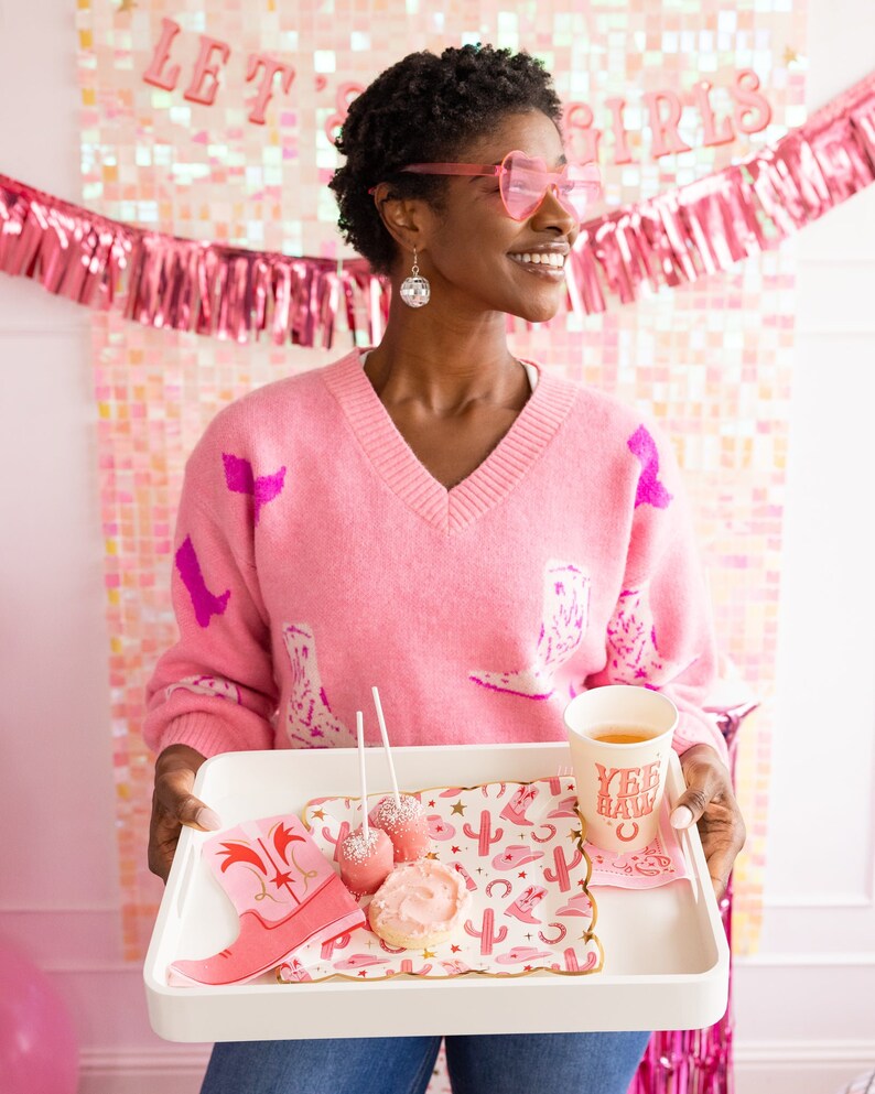 K&ouml;nnte beinhalten: Ein rosa Pullover mit Cowboy-Stiefel-Muster, ein wei&szlig;es Tablett mit rosa und goldenen Servietten, zwei rosa Kuchen am Stiel, ein rosa glasierter Keks und eine rote Tasse mit dem Text "Yee Haw" darauf.
