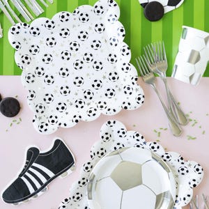 May include: A set of soccer-themed party supplies, including plates, napkins, and a cup. The plates feature a white background with black and silver soccer balls and stars. The napkins feature a black and white soccer shoe. The cup is white with a black and silver soccer ball pattern.
