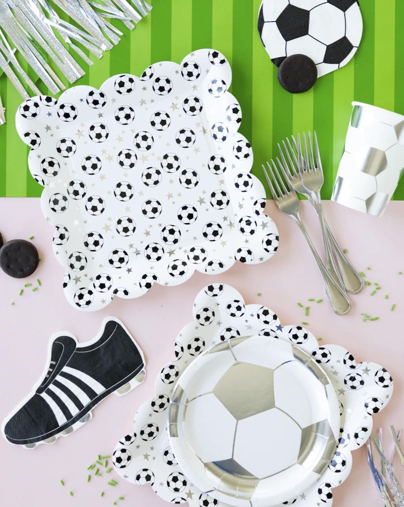 May include: A set of soccer-themed party supplies, including plates, napkins, and a cup. The plates feature a white background with black and silver soccer balls and stars. The napkins feature a black and white soccer shoe. The cup is white with a black and silver soccer ball pattern.