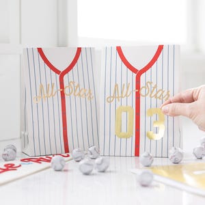 Op de afbeelding: Twee witte en blauwe gestreepte papieren zakken met rode bies en gouden letters die "All-Star" zeggen. Een zak heeft een gouden nummer "03" erop.