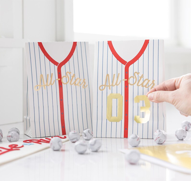 Op de afbeelding: Twee witte en blauwe gestreepte papieren zakken met rode bies en gouden letters die "All-Star" zeggen. Een zak heeft een gouden nummer "03" erop.
