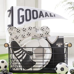 May include: A black wire basket filled with soccer-themed party supplies, including paper plates, a banner that says "GOOAAL!" and a soccer jersey. There are also several black and white soccer balls on a green and white striped surface.