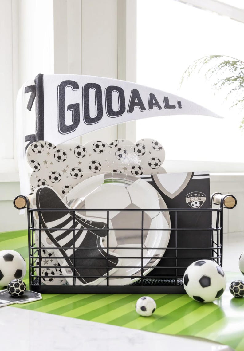 May include: A black wire basket filled with soccer-themed party supplies, including paper plates, a banner that says "GOOAAL!" and a soccer jersey. There are also several black and white soccer balls on a green and white striped surface.