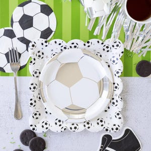 May include: A white and silver soccer ball themed paper plate with a white and silver soccer ball design. The plate is on a white surface with a green and white striped background. There are also chocolate chip cookies, a silver cup, and a black and white soccer shoe.