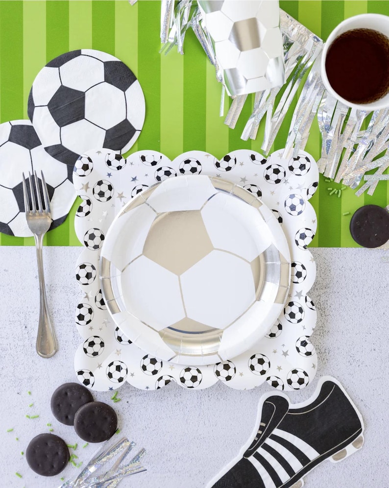 May include: A white and silver soccer ball themed paper plate with a white and silver soccer ball design. The plate is on a white surface with a green and white striped background. There are also chocolate chip cookies, a silver cup, and a black and white soccer shoe.