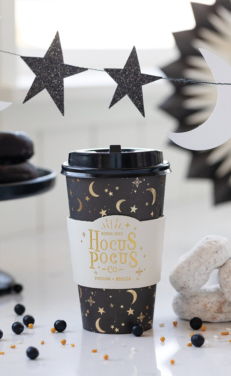 K&ouml;nnte beinhalten: Eine schwarze und goldene Kaffeetasse mit einem Hocus Pocus Co. Logo und einem Mond- und Sternenmuster. Die Tasse steht auf einer wei&szlig;en Oberfl&auml;che mit goldenen und schwarzen Streuseln.