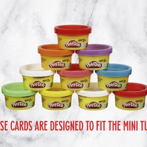 May include: A collection of ten small Play-Doh tubs in various colors, stacked in a pyramid shape. The tubs are yellow with a red, orange, white, green, blue, purple, and pink lid. Each tub has the Play-Doh logo and the text "AGES 2+".