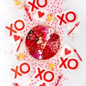 May include: A Valentine's Day table setting with a pink and white polka dot tablecloth. The table is decorated with red and white "XO" plates, red and white heart napkins, and a large bowl of sweets.
