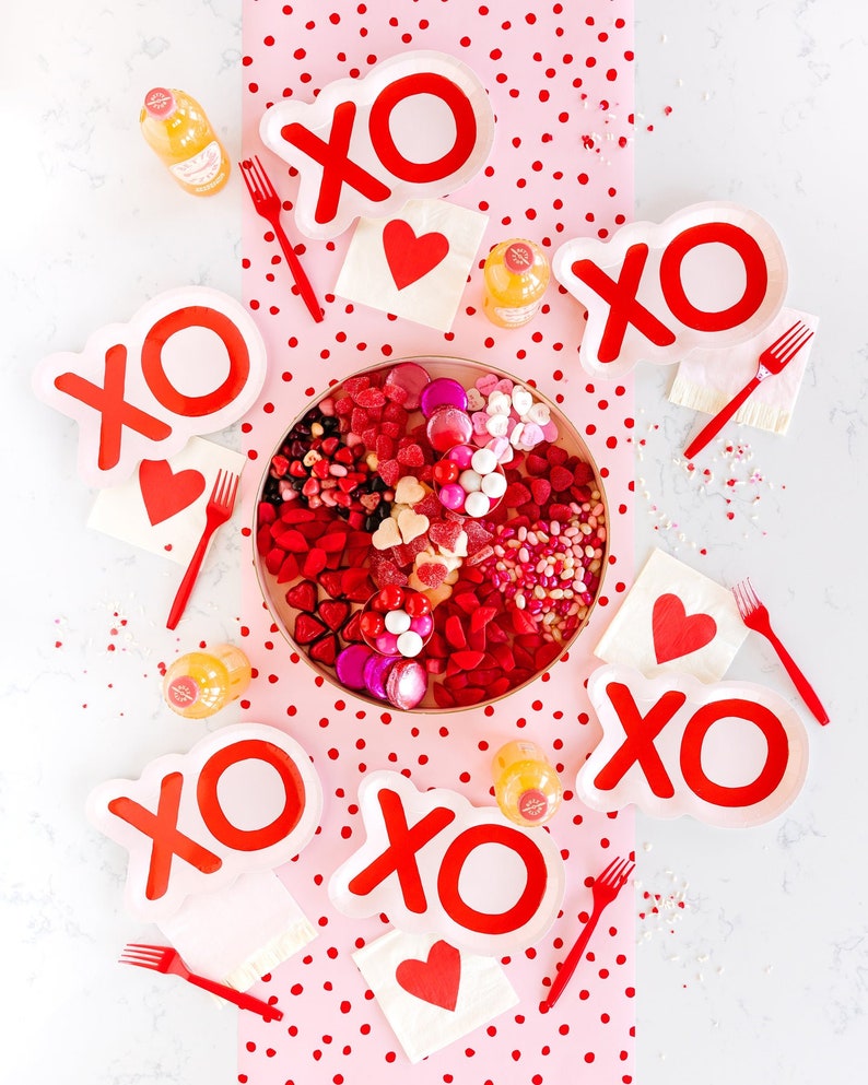 May include: A Valentine's Day table setting with a pink and white polka dot tablecloth. The table is decorated with red and white "XO" plates, red and white heart napkins, and a large bowl of sweets.
