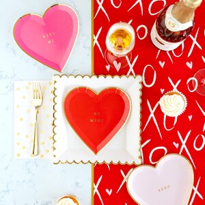 Puede incluir: Una mesa preparada para el D&iacute;a de San Valent&iacute;n con platos de papel en forma de coraz&oacute;n rojo y rosa, tenedores dorados y cupcakes. Los platos tienen las frases "Kiss Me", "Be Mine" y "XOXO" impresas. El mantel es rojo con patrones blancos de "XO" y corazones.