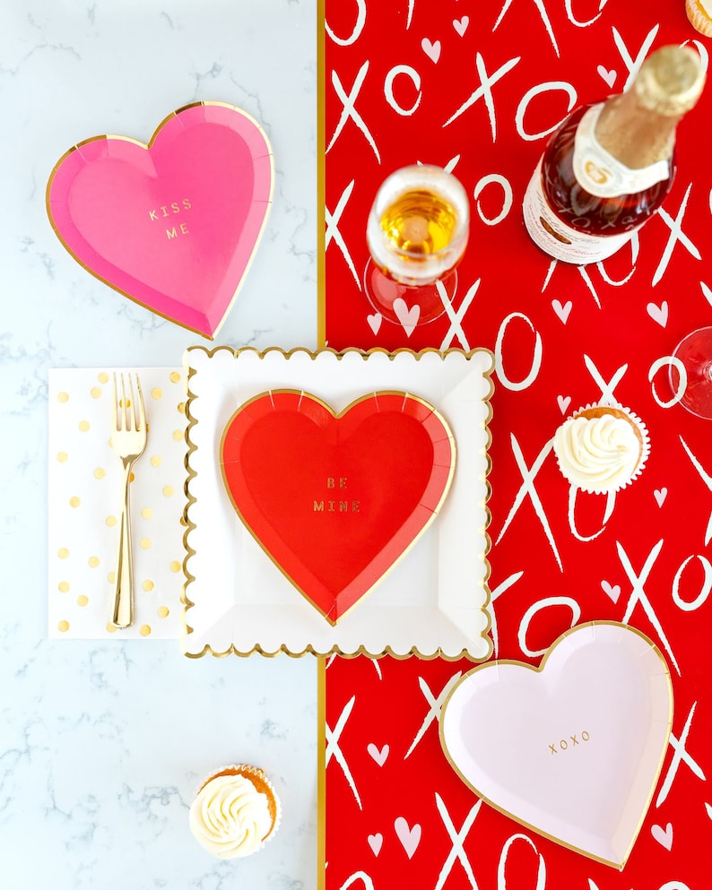 Puede incluir: Una mesa preparada para el D&iacute;a de San Valent&iacute;n con platos de papel en forma de coraz&oacute;n rojo y rosa, tenedores dorados y cupcakes. Los platos tienen las frases "Kiss Me", "Be Mine" y "XOXO" impresas. El mantel es rojo con patrones blancos de "XO" y corazones.