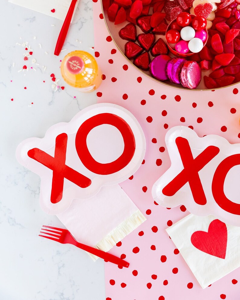 May include: Two pink paper plates with red XO letters on them. The plates are on a pink background with red polka dots. There is a white napkin with a red heart on it and a red fork on the table.