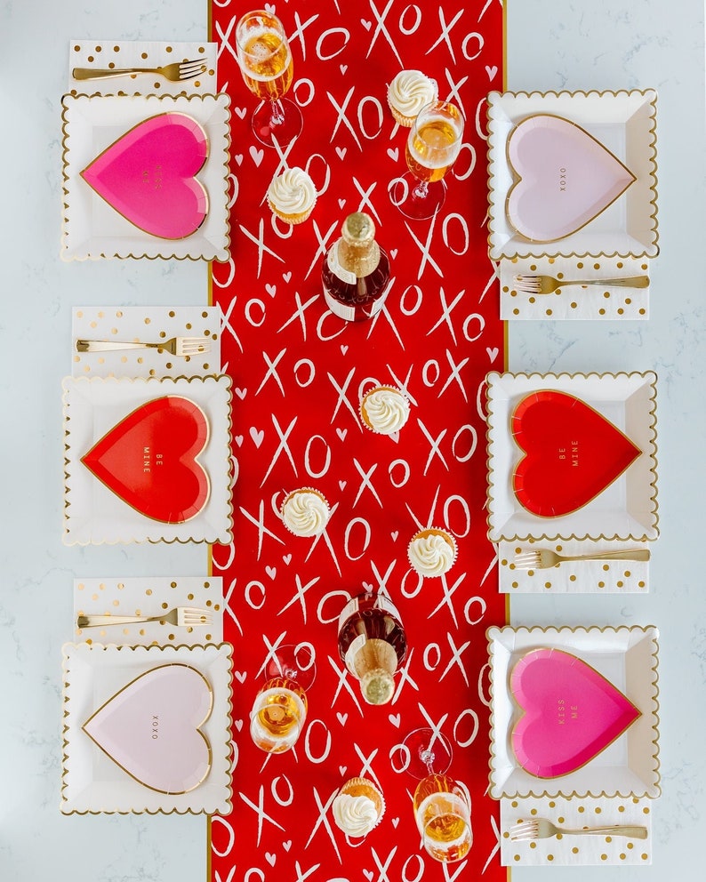 Puede incluir: Un camino de mesa rojo con patrones blancos de "XO" y corazones. Cuatro platos cuadrados blancos con bordes dorados y platos en forma de coraz&oacute;n rosa y rojo con bordes dorados. Los platos tienen el texto "XOXO", "Be Mine", "Kiss Me" y "Be Mine" en ellos. Tambi&eacute;n hay tenedores dorados y servilletas con lunares dorados.