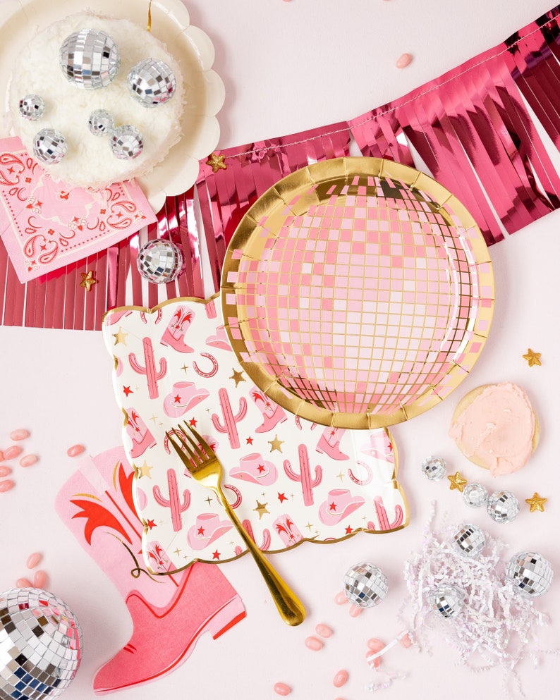 K&ouml;nnte beinhalten: Rosa und goldene Partyteller mit einem Discokugel-Design, rosa und wei&szlig;e Servietten mit einem Kaktus- und Cowboystiefel-Design und silberne Discokugel-Ornamente.