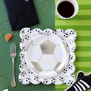 May include: A soccer-themed party table setting with a black and white soccer jersey invitation, a white paper cup with black trim, a white paper plate with a silver soccer ball design, a white paper plate with black soccer balls and stars, and a black and white soccer shoe cutout.
