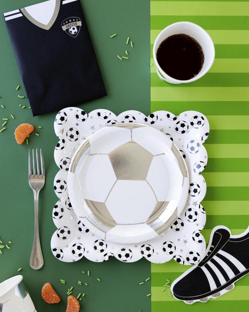 May include: A soccer-themed party table setting with a black and white soccer jersey invitation, a white paper cup with black trim, a white paper plate with a silver soccer ball design, a white paper plate with black soccer balls and stars, and a black and white soccer shoe cutout.