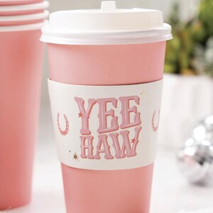 K&ouml;nnte beinhalten: Rosa Einweg-Kaffeetasse mit wei&szlig;em Deckel und einer wei&szlig;en Papiersleeve mit dem Text "Yee Haw" in rosa Buchstaben und goldenen Sternen und Hufeisen-Akzenten.