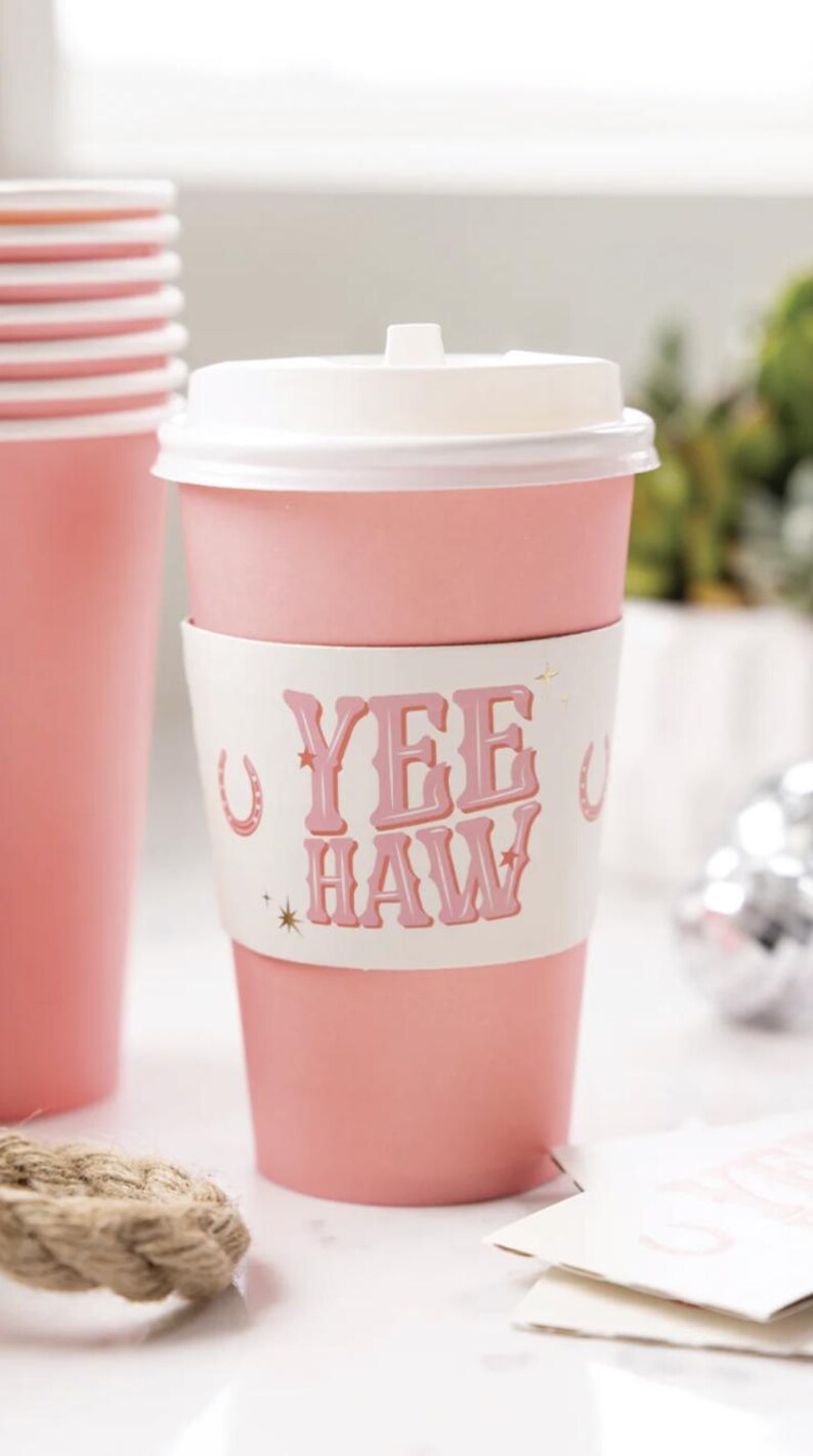K&ouml;nnte beinhalten: Rosa Einweg-Kaffeetasse mit wei&szlig;em Deckel und einer wei&szlig;en Papiersleeve mit dem Text "Yee Haw" in rosa Buchstaben und goldenen Sternen und Hufeisen-Akzenten.