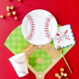 Op de afbeelding: Een baseball-thema tafeldecoratie met een wit papieren bord, een rood-wit papieren bekertje, een gouden vork en een groen-gouden baseballveld-vormig placemat. De placemat heeft een wit servet met de tekst "All-Star Champion" en een baseballknuppel en bal afbeelding. Er zijn ook verspreide karamel popcornkorrels op het rode oppervlak.