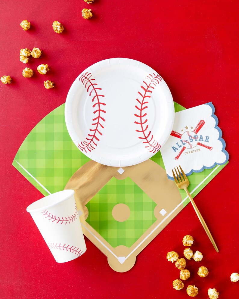 Op de afbeelding: Een baseball-thema tafeldecoratie met een wit papieren bord, een rood-wit papieren bekertje, een gouden vork en een groen-gouden baseballveld-vormig placemat. De placemat heeft een wit servet met de tekst "All-Star Champion" en een baseballknuppel en bal afbeelding. Er zijn ook verspreide karamel popcornkorrels op het rode oppervlak.