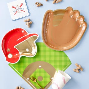 Op de afbeelding: Baseball-thema feestbenodigdheden, waaronder een rood en gouden baseballhelm-vormig papieren bord, een bruin en gouden baseballhandschoen-vormig papieren bord, een wit papieren bekertje met rode baseballnaden, een gouden vork en een groen-wit geblokt papieren tafelkleed met een gouden baseballdiamant.