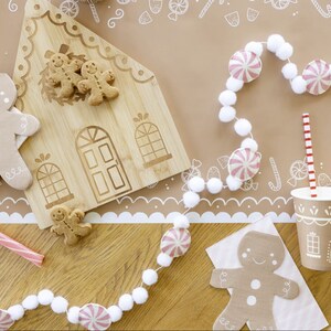 Puede incluir: Una tabla de cortar con forma de casa de jengibre con galletas de jengibre encima. Una guirnalda blanca y rosa con pompones blancos y caramelos de menta rosa cuelga sobre la tabla de cortar. Dos vasos de papel marr&oacute;n con dise&ntilde;os de casas de jengibre y pajitas rojas y blancas rayadas est&aacute;n sobre una mesa marr&oacute;n con un borde estampado blanco y marr&oacute;n.