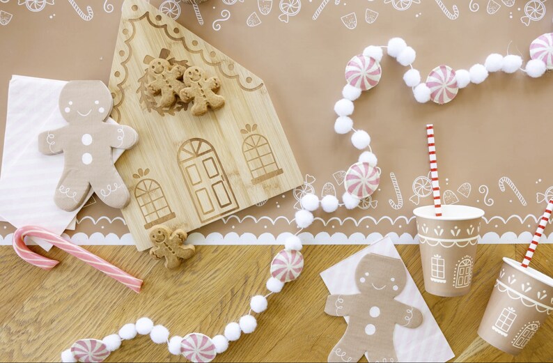 Puede incluir: Una tabla de cortar con forma de casa de jengibre con galletas de jengibre encima. Una guirnalda blanca y rosa con pompones blancos y caramelos de menta rosa cuelga sobre la tabla de cortar. Dos vasos de papel marr&oacute;n con dise&ntilde;os de casas de jengibre y pajitas rojas y blancas rayadas est&aacute;n sobre una mesa marr&oacute;n con un borde estampado blanco y marr&oacute;n.