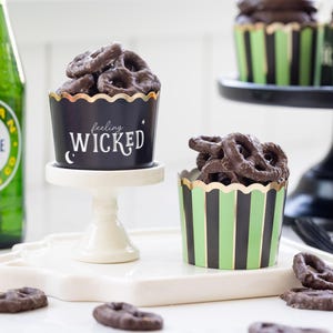 May include: A Halloween-themed image featuring chocolate-covered pretzels in decorative cups. One black cup has gold trim and the words "feeling WICKED". Another cup has green and black stripes. A green glass bottle of ginger ale is visible in the background.