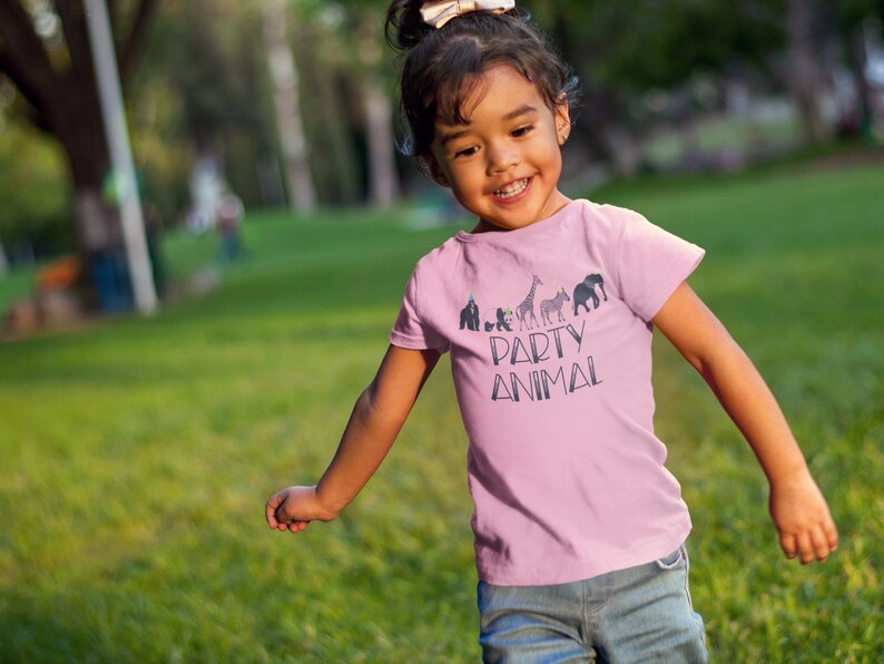 May include: A young girl wearing a pink t-shirt with a graphic of animals wearing party hats and the text "Party Animal". She is smiling and running through a grassy field.