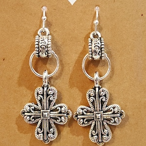 May include: Silver-toned earrings with a cross design. Each earring features a decorative cross pendant with intricate detailing, suspended from a hoop and a top piece with the word "love" engraved. The background is a brown card with the word "Handmade" and a heart.