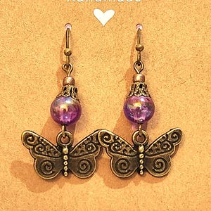 Puede incluir: Pendientes de gota con cuentas de vidrio iridiscente morado y abalorios de mariposa de oro antiguo. Los pendientes están hechos a mano.