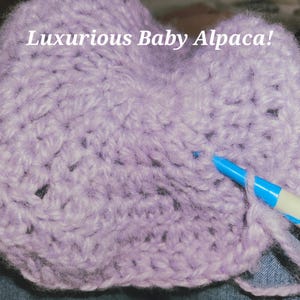 Puede incluir: Primer plano de un artículo de ganchillo en un suave color lavanda. El texto "Luxurious Baby Alpaca!" es visible en la parte superior. Un bolígrafo azul y blanco está insertado en el trabajo de ganchillo.