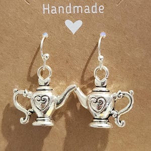 May include: A pair of silver-colored earrings shaped like genie lamps. Each lamp has a heart design and a curved spout. The earrings hang from silver hooks. The background is a brown card with the word "Handmade" and a white heart.