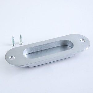 May include: A brushed silver recessed pull with an oval finger hole, accompanied by two silver screws. The hardware is designed for installation in a door or drawer, providing a sleek, modern aesthetic.