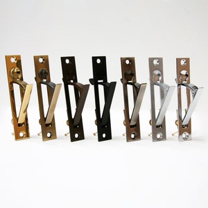 May include: A collection of eight different metal flush pulls in various finishes, including gold, brass, bronze, black, and silver. Each pull has a rectangular backplate and a V-shaped handle. The pulls are arranged in a row against a white background.