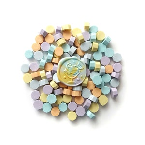 May include: A collection of pastel-coloured wax seals in various shades of blue, yellow, and purple. The wax seals are hexagonal in shape and are arranged in a circular pattern. A single yellow wax seal with a design of a cocktail glass with fruit is in the centre.