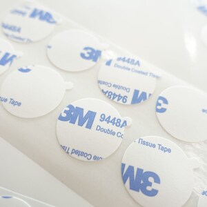 Peut inclure: Gros plan sur plusieurs autocollants adh&eacute;sifs ronds, blancs, avec la marque 3M bleue et le texte "9448A Double Coated Tape".