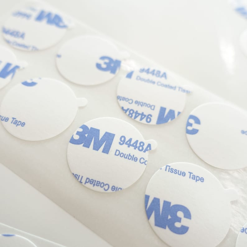 Peut inclure: Gros plan sur plusieurs autocollants adh&eacute;sifs ronds, blancs, avec la marque 3M bleue et le texte "9448A Double Coated Tape".