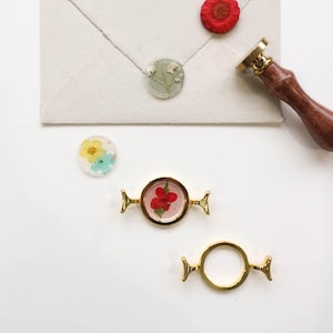 May include: An assortment of decorative wax seals and a wax stamp. The seals feature dried flowers encased in clear resin. One seal is on an envelope. Two gold-colored metal clasps are also present, one with a red flower.