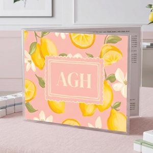May include: A clear acrylic menu holder with a pink background featuring a lemon and floral pattern. The holder has the letters "AGH" in a pink frame. Mahjong tiles and a lemon branch in a vase are also visible.