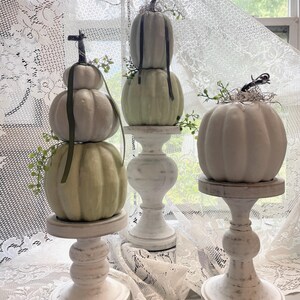 May include: Three white and sage green decorative pumpkins stacked on white distressed wooden pedestals. The pumpkins are accented with dark green ribbons, small sprigs of greenery, and wispy dried fibers attached to their dark brown stems.