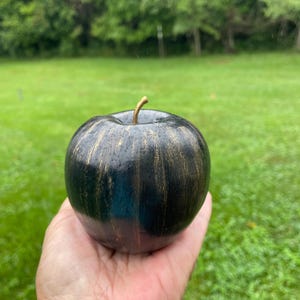 May include: A decorative apple sculpture featuring a smooth, rounded black surface with subtle, vertical streaks of metallic gold paint. The object includes a thin, curved gold-colored stem extending from the top center of the fruit-shaped form.