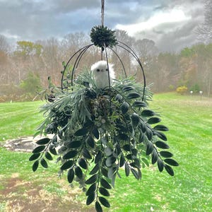 May include: A white bird figurine sits in a nest inside a black wire sphere hanging from a ceiling. The sphere is surrounded by green leafy branches.