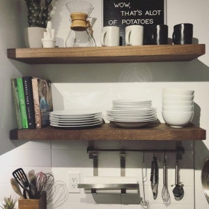 May include: A kitchen interior with two wooden shelves. The top shelf features white and black mugs, a glass coffee maker, and a sign saying "Wow, that's a lot of potatoes." The lower shelf displays stacks of white plates and bowls, and cookbooks.