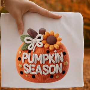 May include: An orange pumpkin-shaped decoration with the words "PUMPKIN SEASON" in white. The pumpkin has a knitted texture and is adorned with a sunflower, bow, and leaves. Black dots and gold stars decorate the pumpkin.