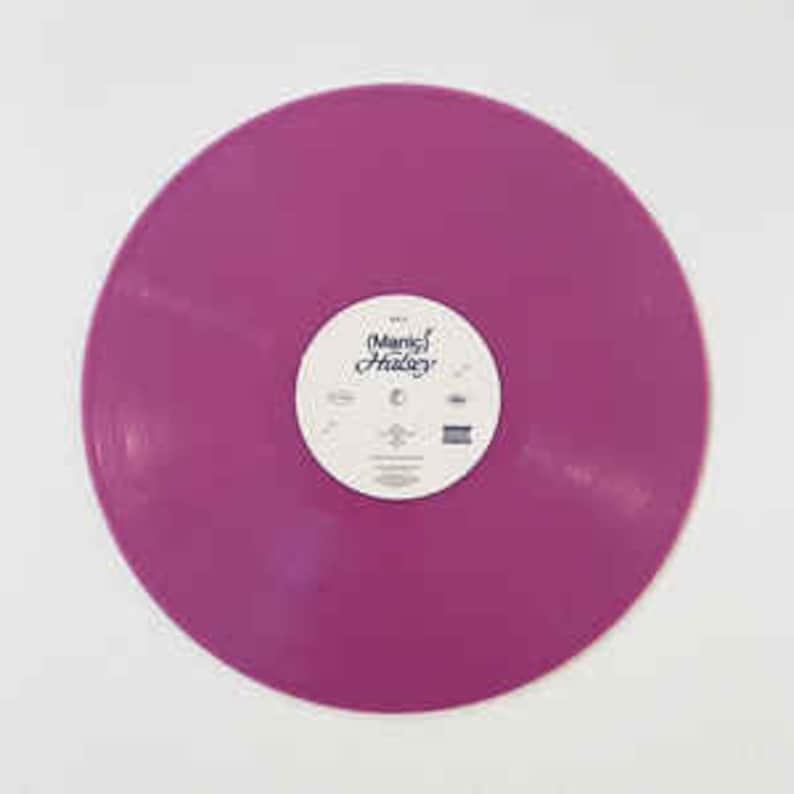 Puede incluir: Un disco de vinilo morado liso con una etiqueta blanca en el centro. La etiqueta presenta el texto "Manic Halsey" y otros textos y s&iacute;mbolos peque&ntilde;os. El disco es circular y est&aacute; sobre una superficie blanca.