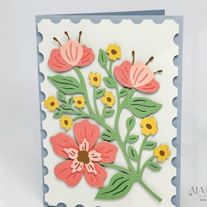 May include: A white card with a blue scalloped border features a paper craft design of pink and yellow flowers with green stems and leaves.