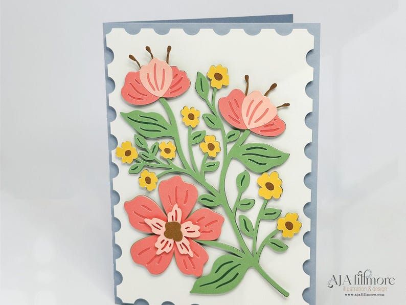 May include: A white card with a blue scalloped border features a paper craft design of pink and yellow flowers with green stems and leaves.
